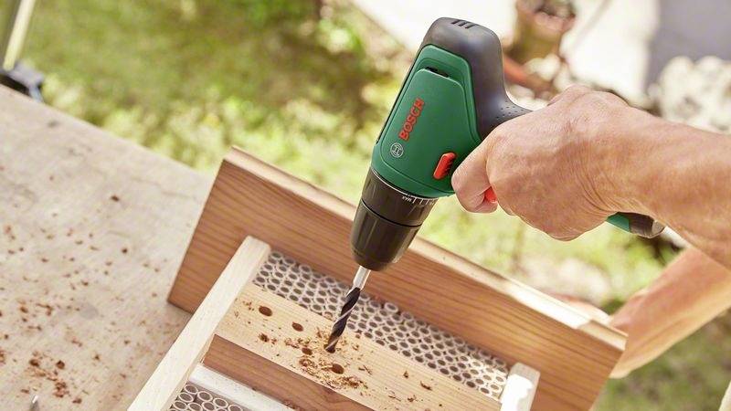 Eine Person bohrt ein Loch in ein Holzstück mit einem grünen Akku-Bohrschrauber. Holzspäne sind auf der Werkbank sichtbar.