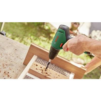 Eine Person bohrt ein Loch in ein Holzstück mit einem grünen Akku-Bohrschrauber. Holzspäne sind auf der Werkbank sichtbar.
