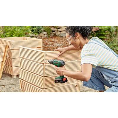 Eine Person mit Schutzbrille verwendet eine Bohrmaschine, um einen Holzkasten im Freien zusammenzubauen. Pflanzen und Steine im Hintergrund.