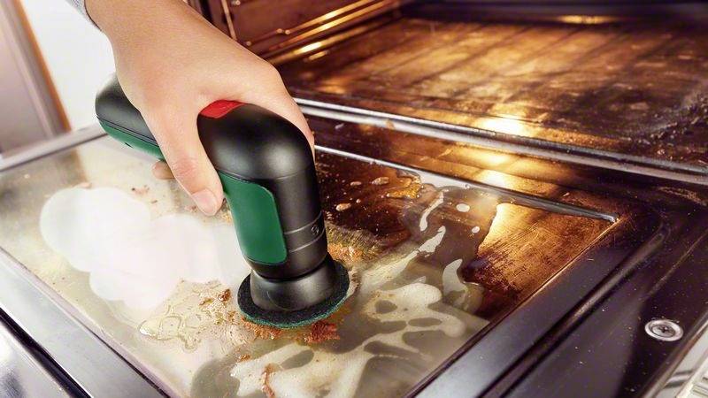 Eine Hand reinigt eine verschmutzte Ofentür mit einem elektrischen Reinigungspad, um eingebrannten Schmutz zu entfernen.