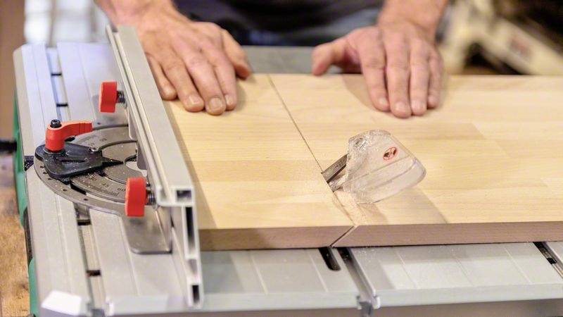 Eine Person schneidet ein Holzbrett präzise mit einer Tischkreissäge. Hände führen das Holz, während die Säge durch die Mitte schneidet.