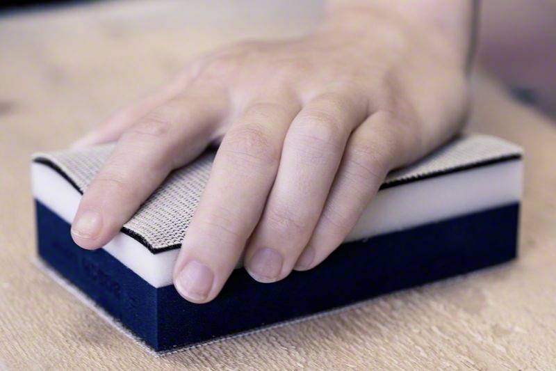 Eine Hand hält einen Reinigungsschwamm auf einer Holzoberfläche, bereit zum Säubern oder Schleifen.