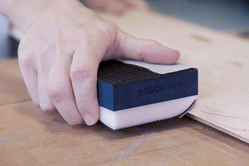 Eine Hand schleift Holz mit einem Schleifblock der Marke Bosch. Der Block hat eine schwarze Schleiffläche und breitet sich Staub auf der Werkbank aus.