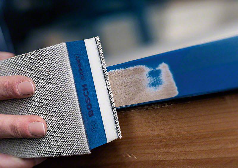 Eine Hand schleift mit einem Schleifblock einen blauen Holzrahmen, um eine Holzfläche freizulegen. Bosch-Logo auf dem Schleifblock sichtbar.