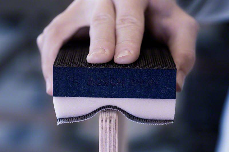 Hand hält Schleifblock über eine Kante eines Holzstücks. Schleifpapier an der Unterseite zeigt Gebrauchsspuren.