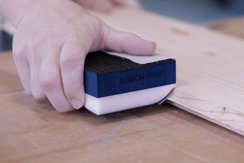 Eine Hand hält einen Bosch-Schleifblock, um eine Holzkante zu glätten. Der Schleifprozess erzeugt Holzstaub auf der Oberfläche.