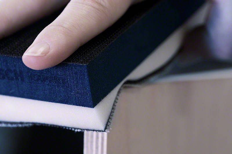 Eine Hand hält einen blauen Schleifblock, der auf einem weißen Schaumstoff liegt und eine glatte Kante bearbeitet.
