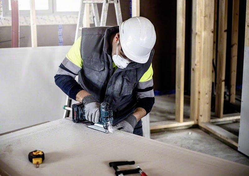 Eine Person mit Schutzhelm und Maske arbeitet mit einem Bohrer an einer Holzplatte in einem unfertigen Raum mit Leitern und Holzrahmen.