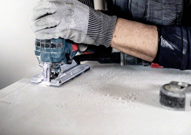 Arbeiter sägt mit einer elektrischen Stichsäge ein Brett, trägt dabei graue Arbeitshandschuhe. Holzstaub auf der Arbeitsfläche sichtbar.