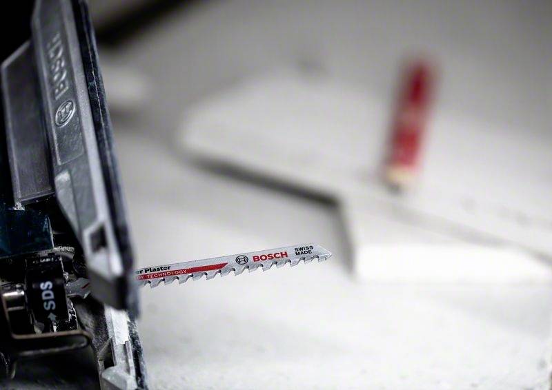 Eine Stichsäge mit einem Bosch-Sägeblatt ist bereit zum Schneiden. Im Hintergrund liegen Werkzeuge unscharf auf einem Tisch.