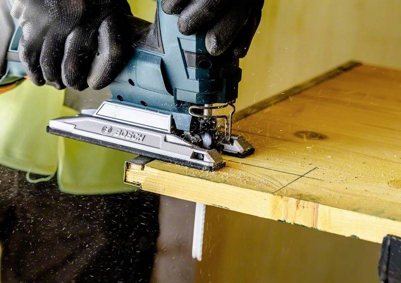Eine Person schneidet mit einer elektrischen Stichsäge ein Stück Holz entlang einer gezeichneten Linie ab. Sägespäne fliegen umher.