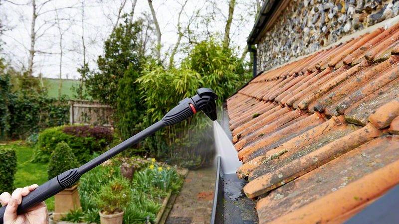 Eine Person reinigt mit einem Hochdruckreiniger ein schräges Ziegeldach, umgeben von einem grünen Garten.