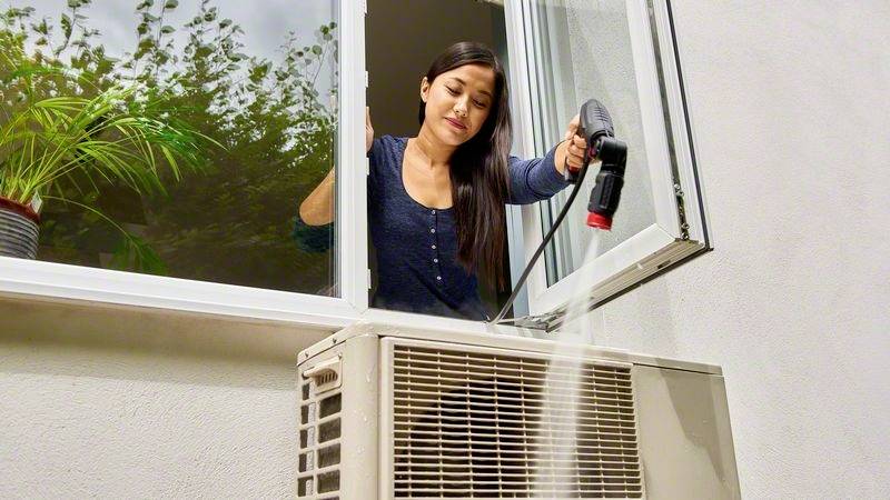 Frau reinigt Außeneinheit einer Klimaanlage mit Wassersprühgerät durch ein geöffnetes Fenster, Pflanze im Hintergrund.