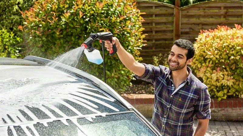 Ein lächelnder Mann wäscht das Dach eines Autos mit einem Wasserstrahl und Reinigungsmittel im Freien an einem sonnigen Tag.