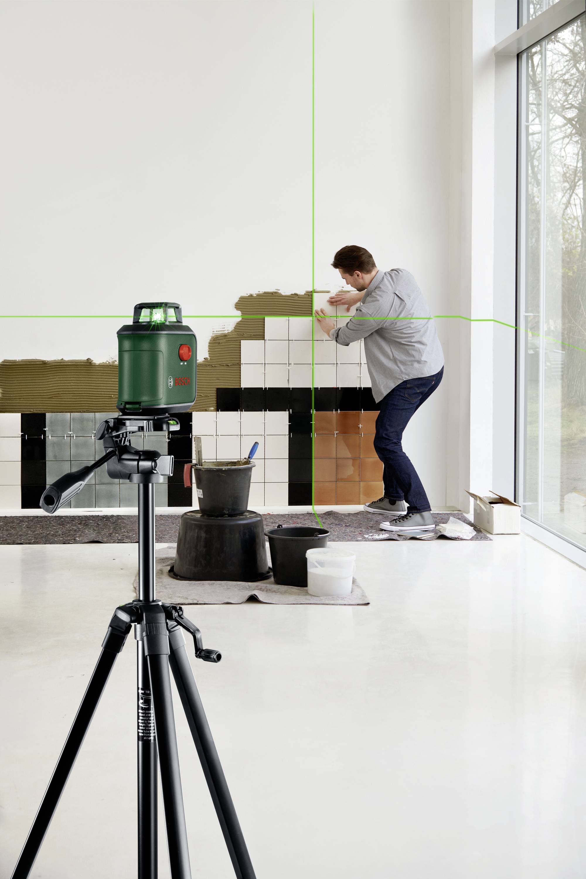 Ein Mann setzt schwarze und weiße Fliesen an eine Wand. Ein grüner Laser-Nivellierer auf einem Stativ projiziert Linien zur Ausrichtung.