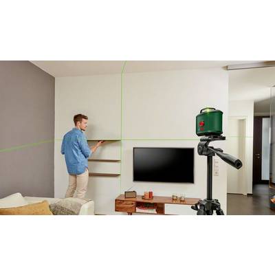 Ein Mann montiert ein Regal an einer Wohnzimmerwand mit Hilfe eines grünen Laser-Nivelliergeräts auf einem Stativ.