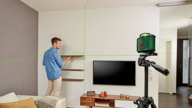 Ein Mann richtet ein Regal mit Hilfe eines grünen Laser-Nivelliergeräts aus, das auf einem Stativ in einem Wohnzimmer steht.