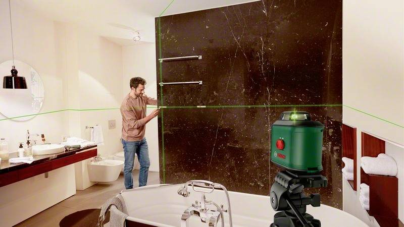 Ein Mann richtet mit einem Lasergerät eine grüne Linie an einer Badezimmerwand aus. Im Hintergrund sind eine Badewanne und ein Waschbecken.