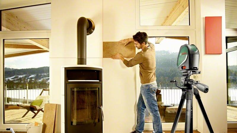 Ein Mann befestigt ein rechteckiges Stück Holz an einer Wand neben einem Ofen. Ein Lasergerät auf einem Stativ zeigt die Ausrichtung an.