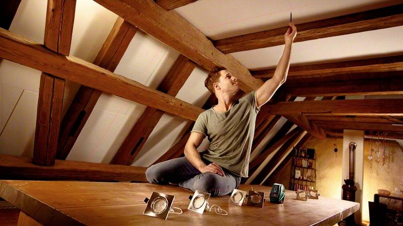 Eine Person sitzt auf einem Holztisch in einem Dachzimmer und hält ein Handy hoch. Vier Lampen sind vor ihr auf dem Tisch platziert.