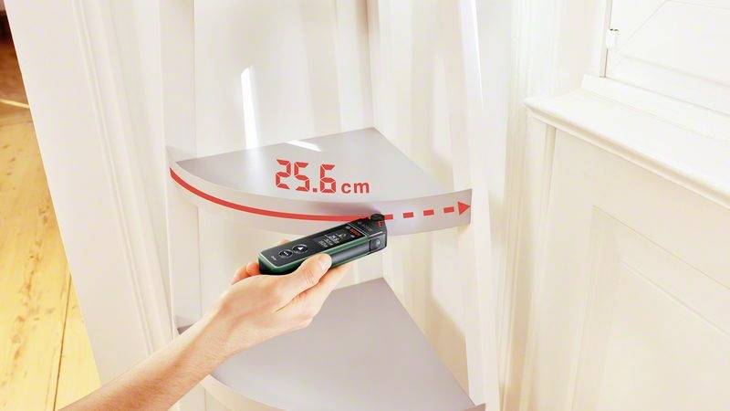 Ein Handgerät misst den Abstand zu einer Wand und zeigt '25,6 cm' an. Roter Laserstrahl markiert die Distanz auf der Wand.