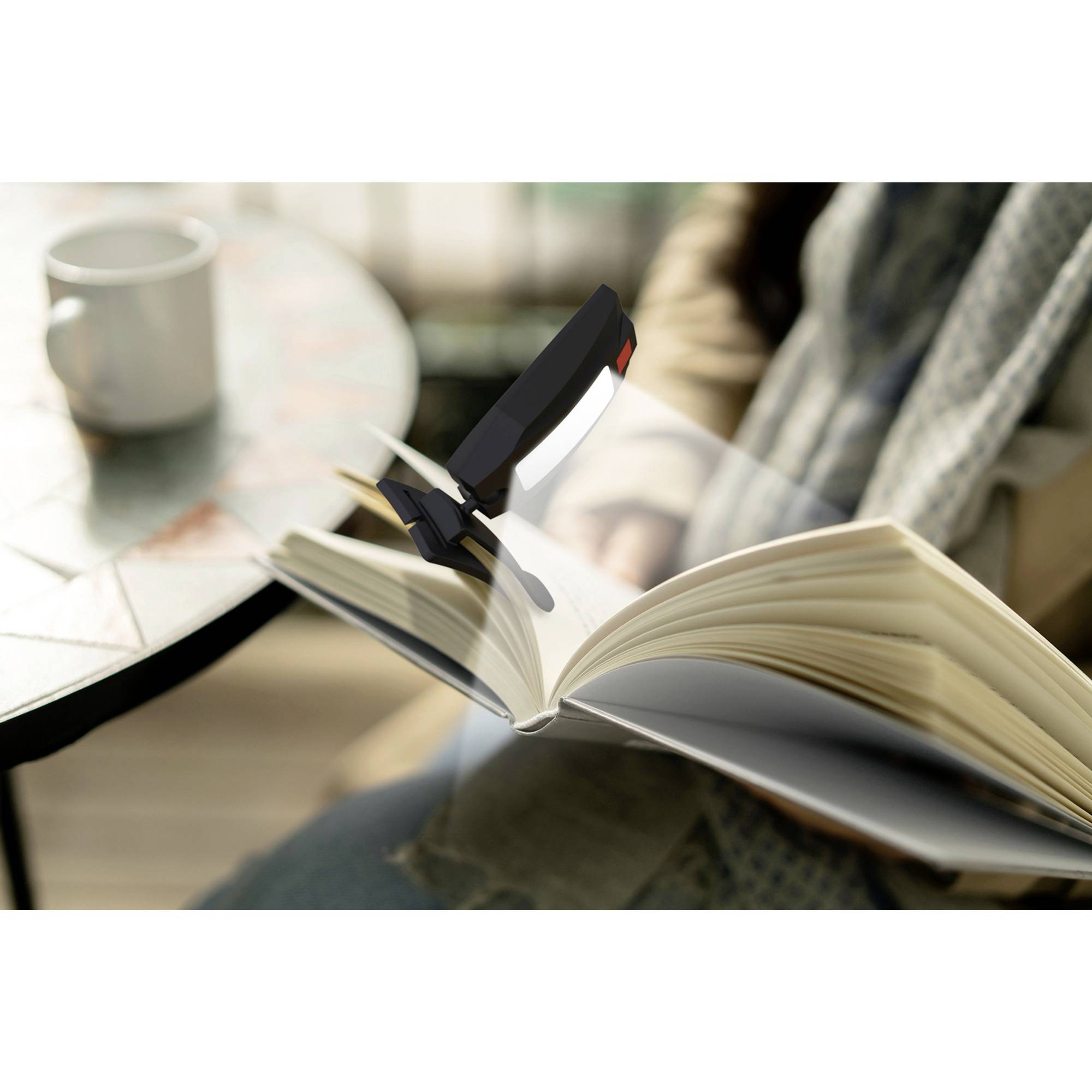 Eine aufgeschlagene Buchseite wird von einer angebrachten Leselampe beleuchtet. Im Hintergrund steht eine weiße Tasse auf einem Tisch.