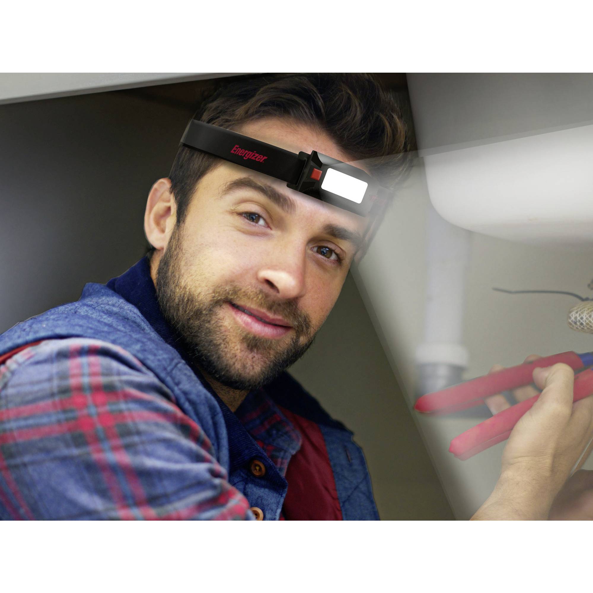 Mann trägt Stirnlampe, arbeitet an Rohr unter Spüle, hält Zange in der Hand. Focus auf Reparaturarbeit mit hellem Licht.