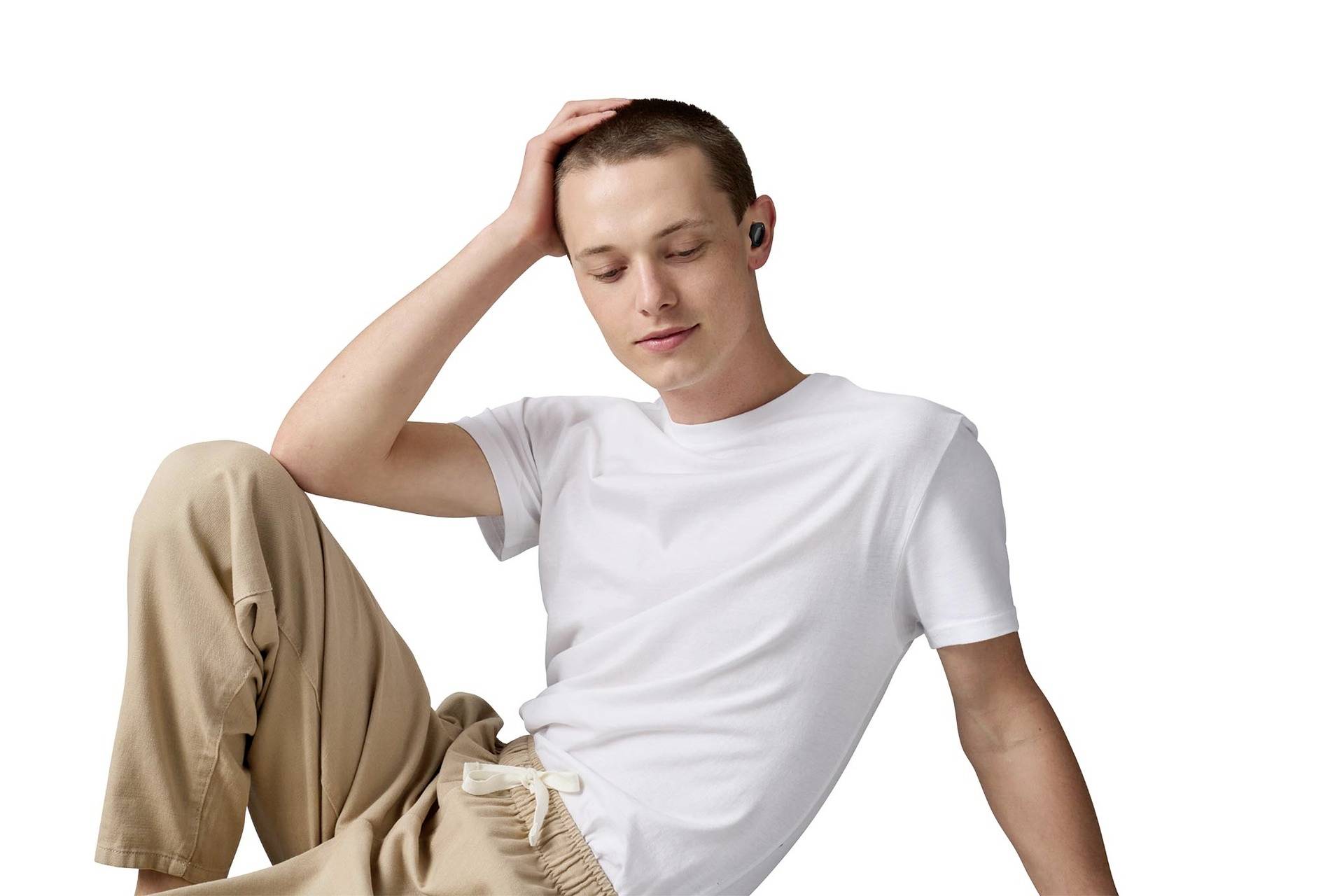 Eine Person sitzt entspannt, trägt eine weiße T-Shirt und beige Hose, hört mit kabellosen Ohrhörern Musik und lächelt leicht.