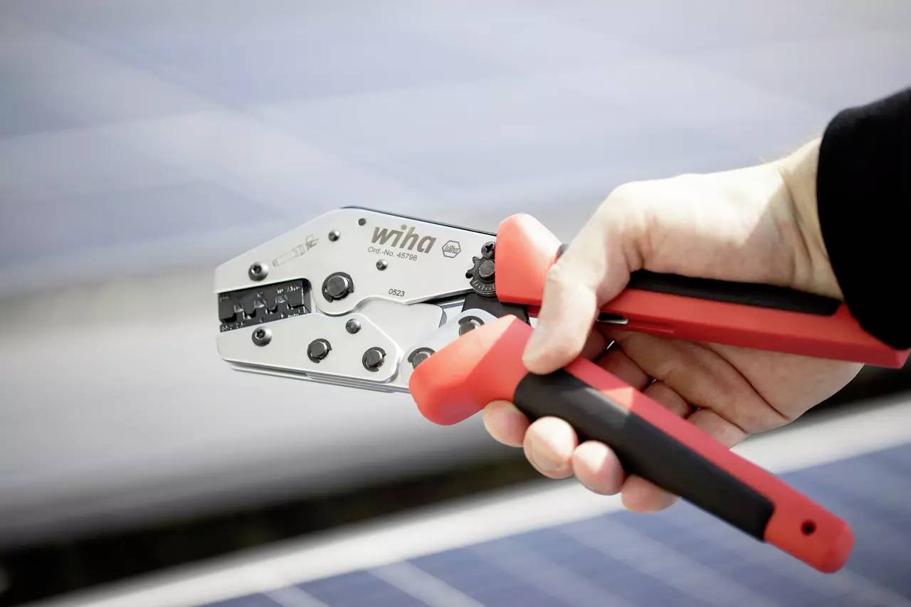 Eine Hand hält eine Crimpzange mit rotem Griff, im Hintergrund unscharfe Solarzellen. Werkzeug von Wiha zum Verbinden von Kabeln.