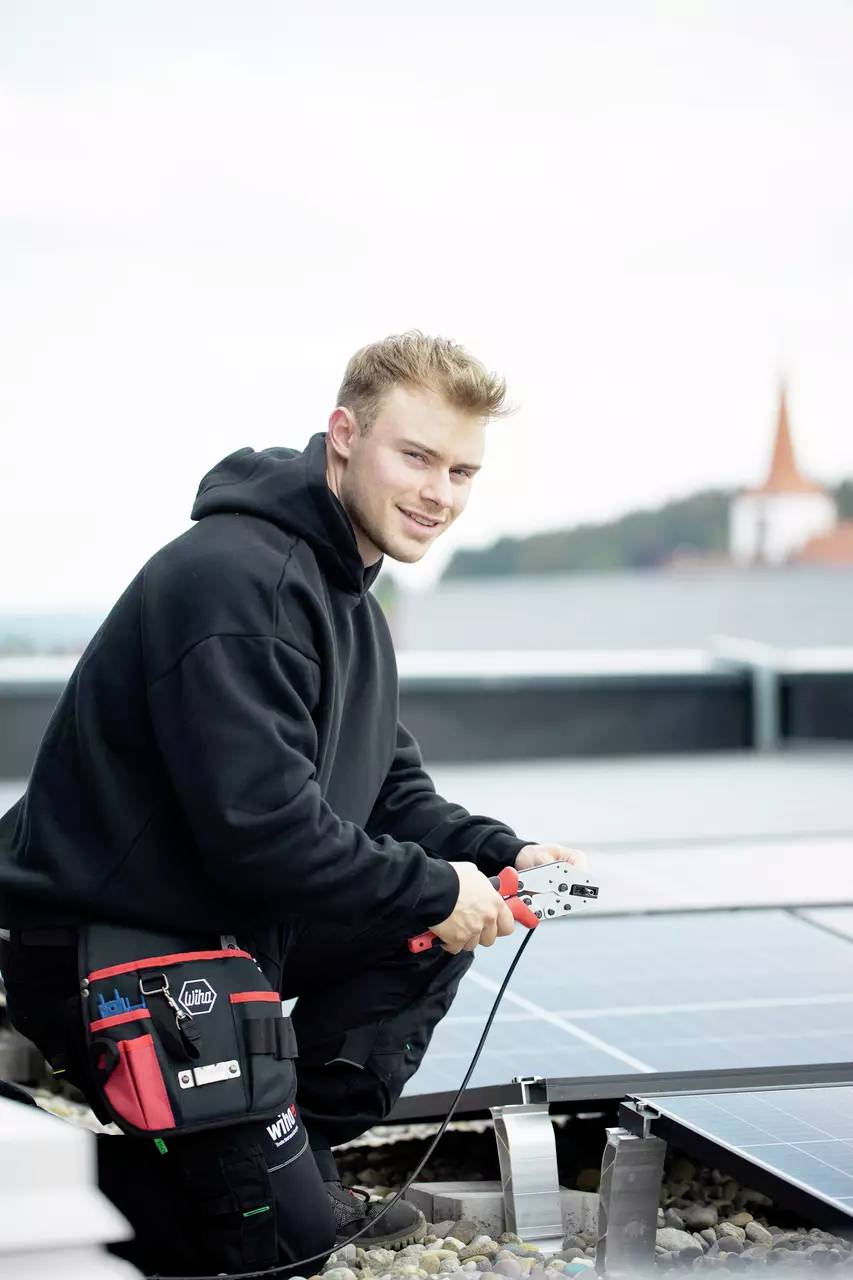 Eine Person in Arbeitskleidung arbeitet an Solarmodulen auf einem Dach. Im Hintergrund ist eine verschwommene Kirche zu sehen.