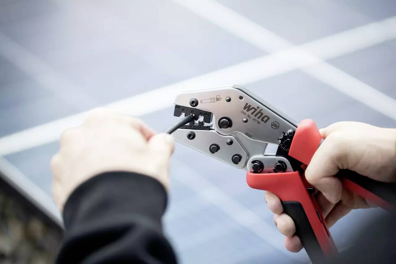 Eine Person verwendet eine Crimpzange, um ein Kabel zu bearbeiten. Im Hintergrund sind Solarmodule zu sehen.