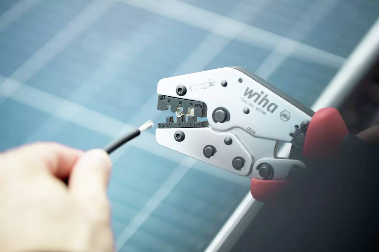 Eine Hand hält eine Crimpzange der Marke Wiha, die ein Kabel bearbeitet. Im Hintergrund sind unscharfe Solarzellen zu sehen.