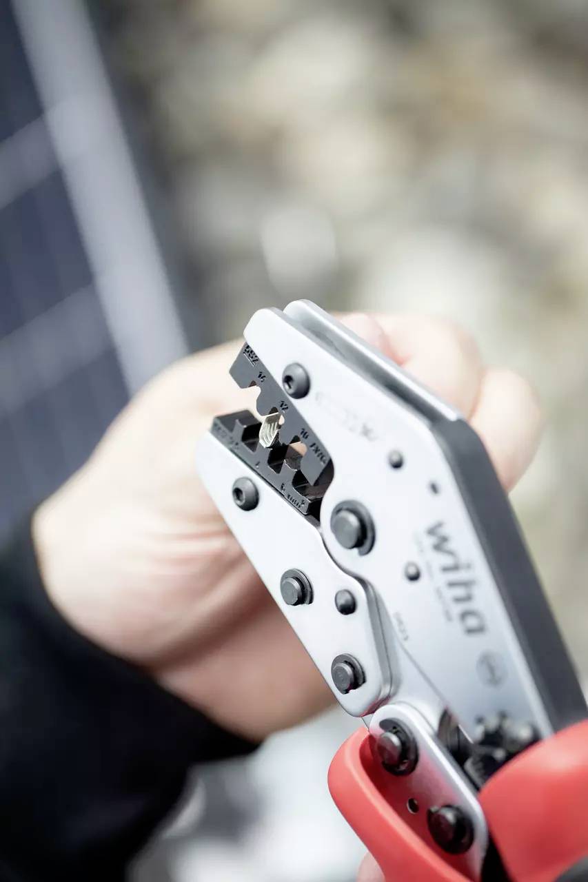 Hand hält eine Crimpzange, die ein Kabel bearbeitet. Im Hintergrund unscharf. Umgebungsdetails sind nicht erkennbar.