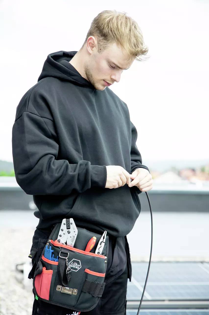 Ein junger Mann in einem schwarzen Kapuzenpullover arbeitet mit Kabeln. Er trägt einen Werkzeuggürtel und steht im Freien.