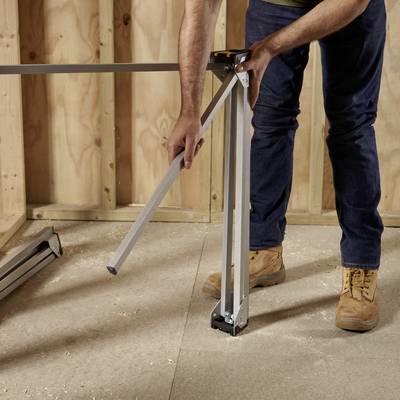 'Person justiert die Höhe eines Tischbeins mit Werkzeug, während sie in einem unvollständigen Raum mit Holzverkleidung steht.'