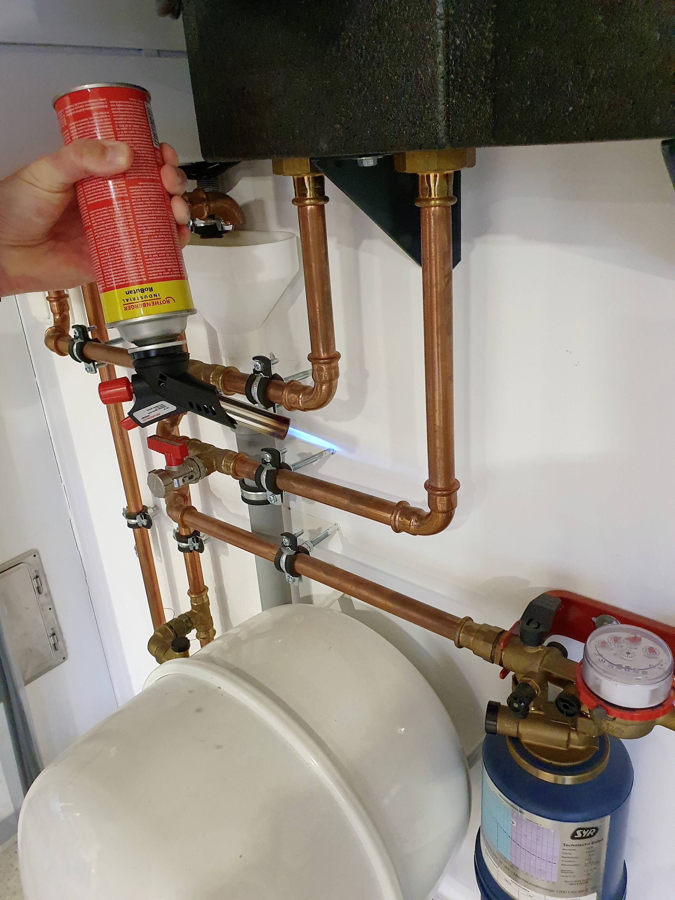 Kupferrohre und Armaturen für Sanitärinstallationen, mit einem Druckmesser und einer Person, die eine Spraydose hält, sichtbar.