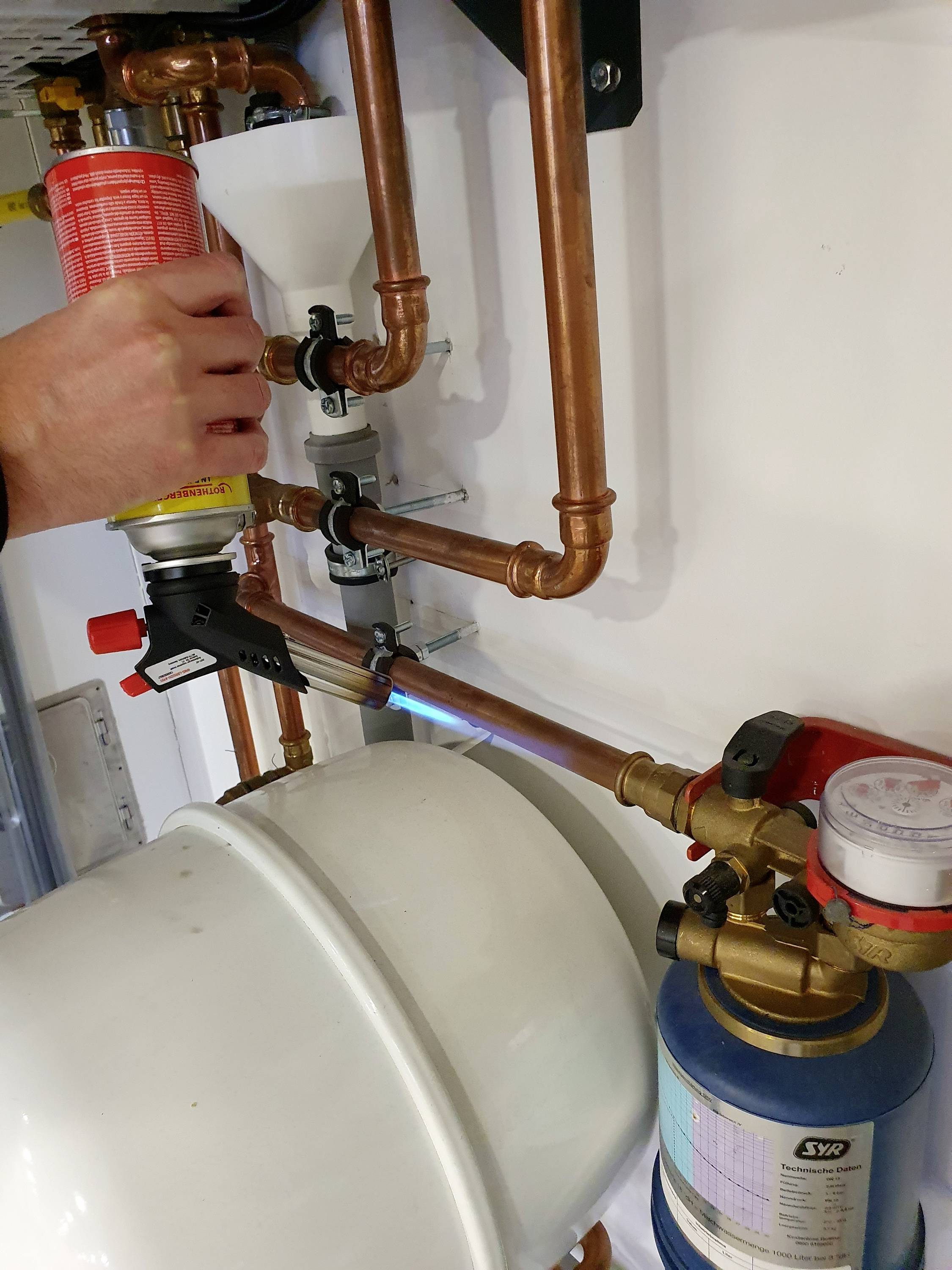 Eine Person verwendet eine Gasfackel, um Kupferrohre an einem Heizungssystem zu verlöten. Ein Druckbehälter ist im Vordergrund sichtbar.