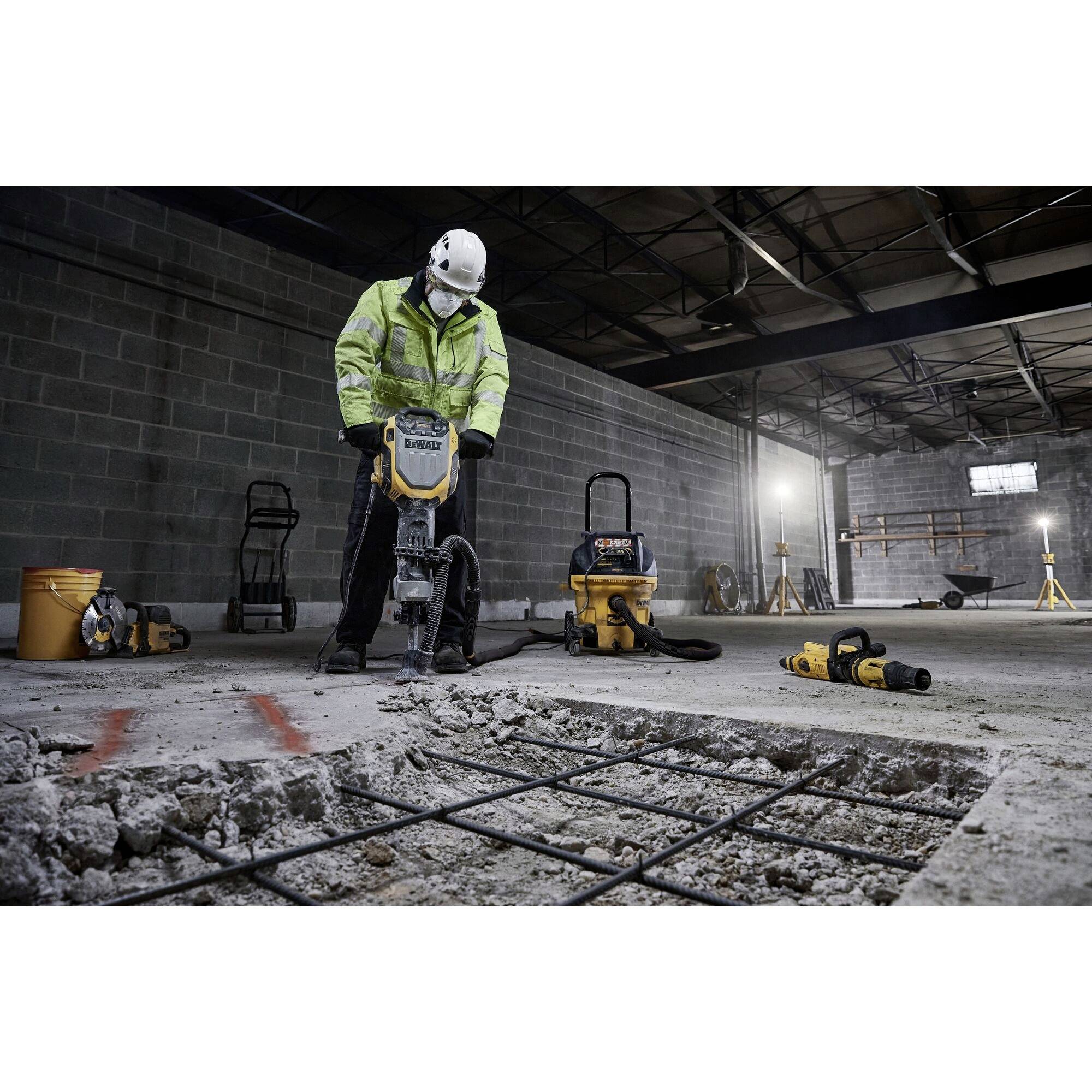 Eine Person in Arbeitskleidung benutzt einen Presslufthammer auf einer Baustelle. Im Hintergrund stehen leere Handwagen und Baugeräte.