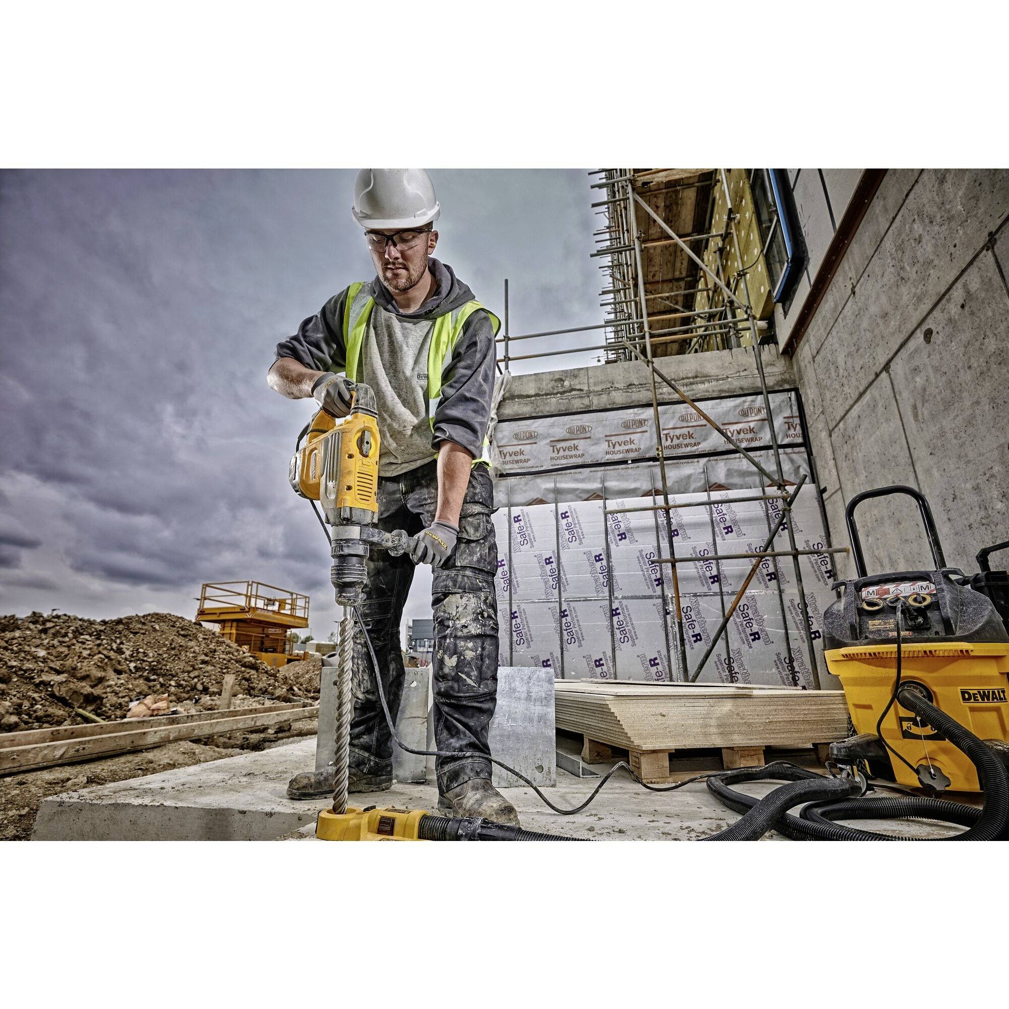 Ein Bauarbeiter benutzt eine Bohrmaschine auf einer Baustelle, umgeben von Gerüsten und Baumaterialien. Er trägt Schutzkleidung und einen Helm.