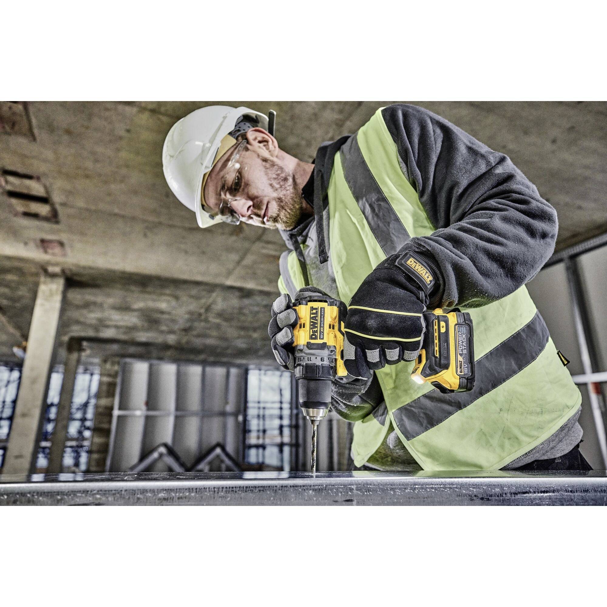 Ein Bauarbeiter mit Schutzhelm und Weste benutzt eine gelbe Bohrmaschine auf einer Baustelle. Im Hintergrund sind Betonwände zu sehen.