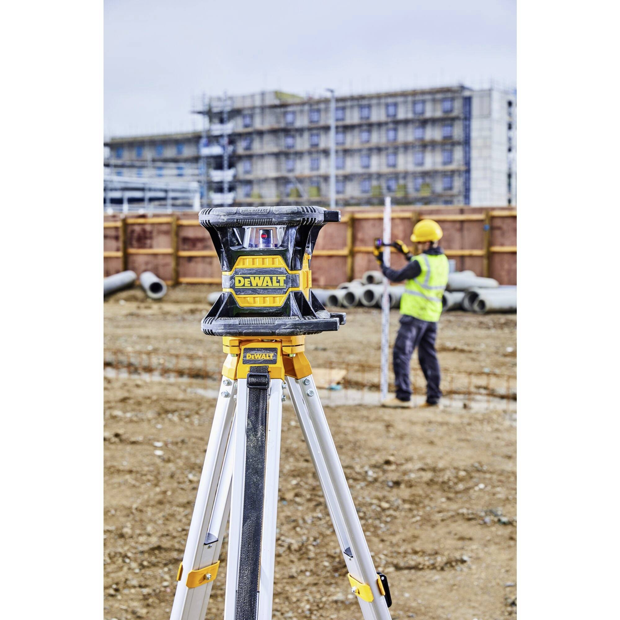'Baustelle mit einem Stativ und Laser-Nivelliergerät im Vordergrund; Bauarbeiter im Hintergrund misst Bodenhöhe vor einem Baugerüst.'