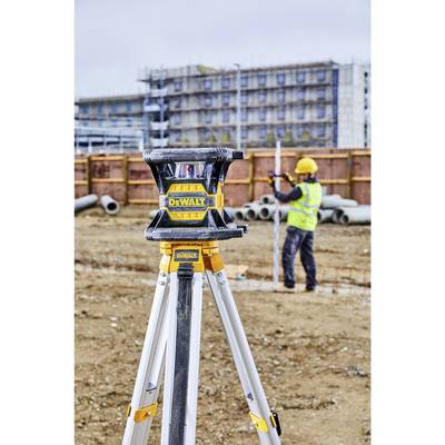 'Baustelle mit einem Stativ und Laser-Nivelliergerät im Vordergrund; Bauarbeiter im Hintergrund misst Bodenhöhe vor einem Baugerüst.'