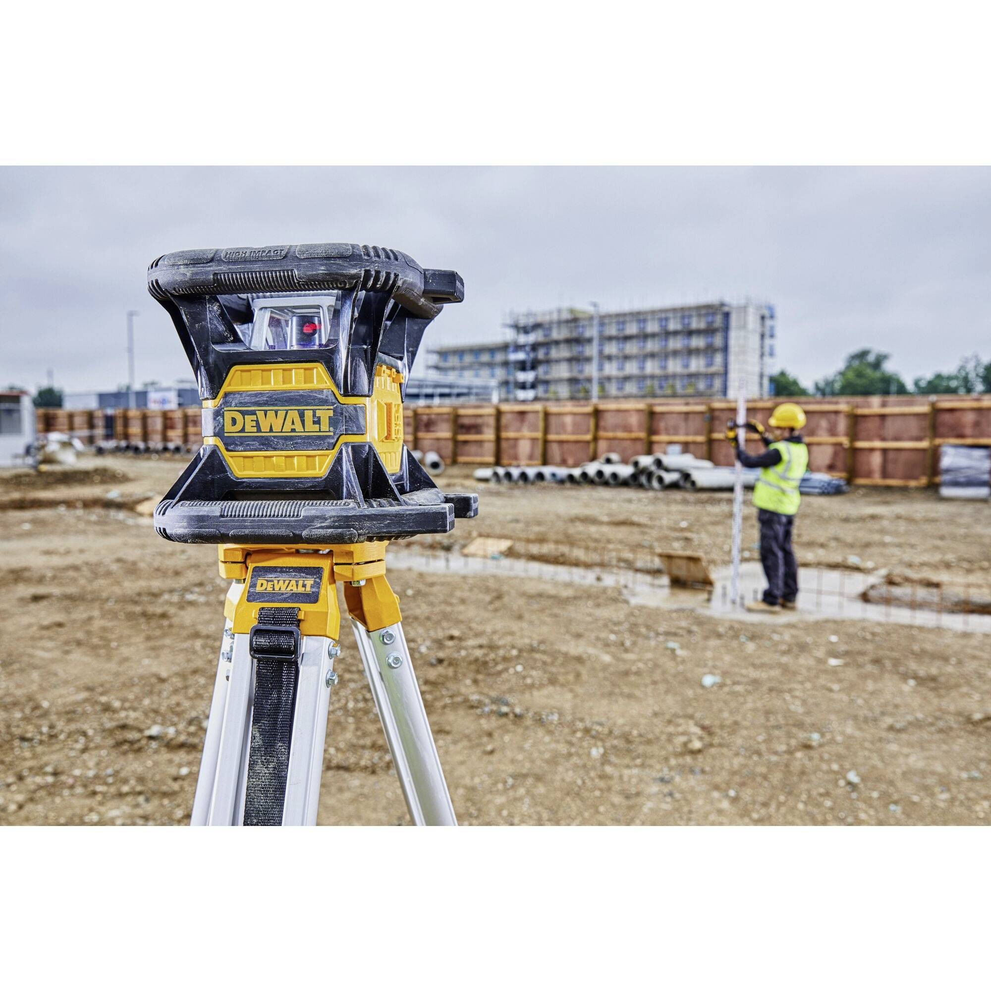 'Baustellenszene mit einem Laser-Nivelliergerät auf einem Stativ im Vordergrund. Ein Bauarbeiter führt Vermessungen im Hintergrund durch.'