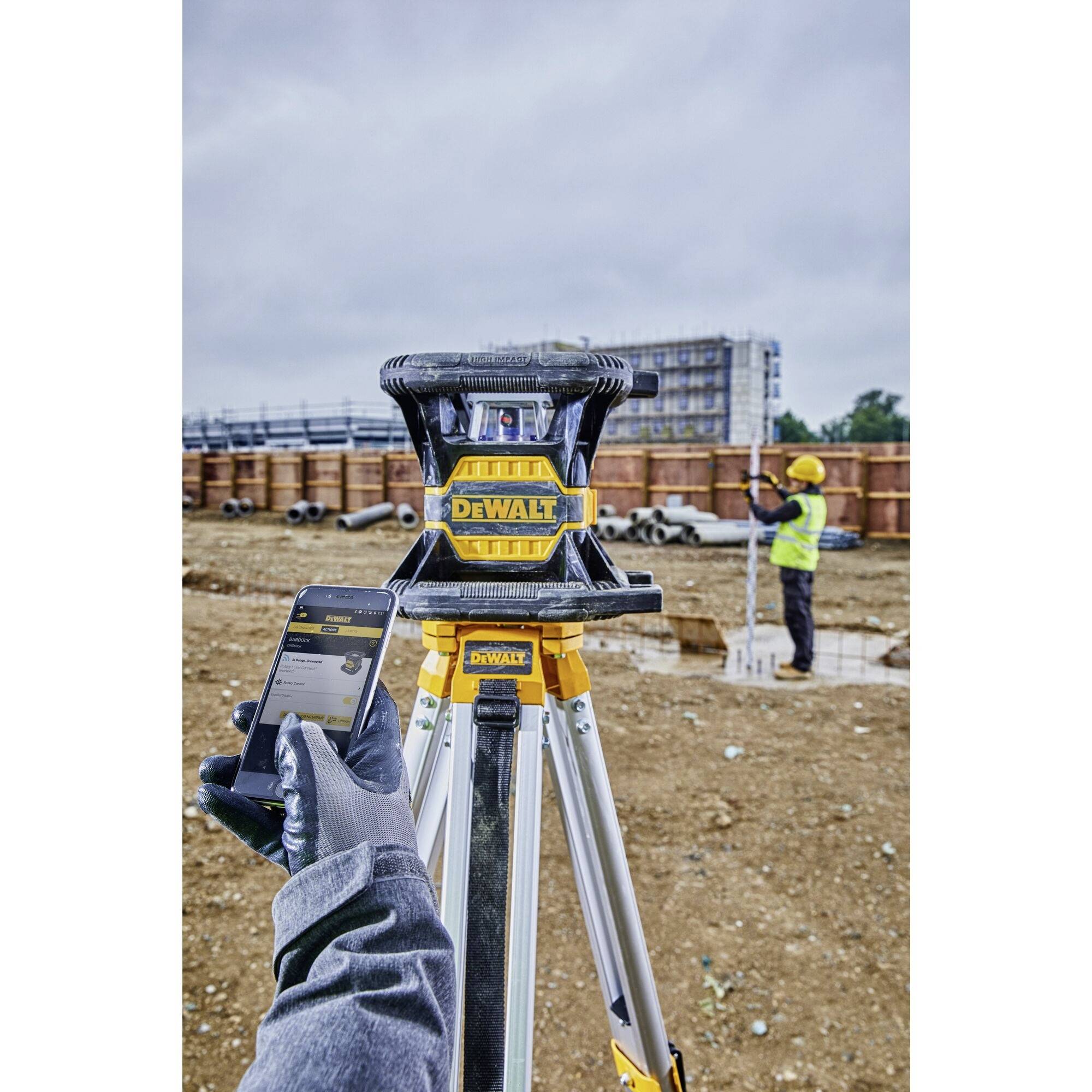 'Arbeiter auf Baustelle mit gelbem Helm im Hintergrund. Im Vordergrund ein Vermessungsgerät und ein Smartphone in einer Hand.'
