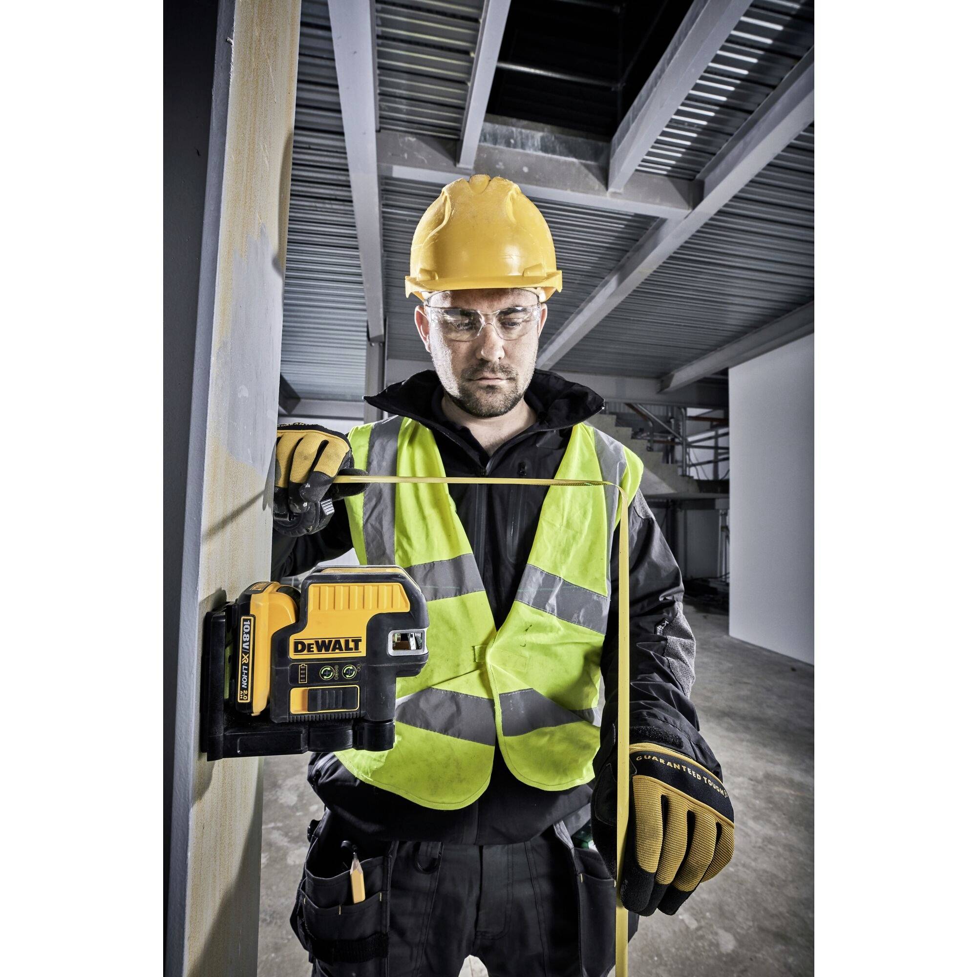 Ein Bauarbeiter mit Helm und Warnweste misst mit einem Lasergerät die Höhe einer Wand in einem industriellen Gebäude.