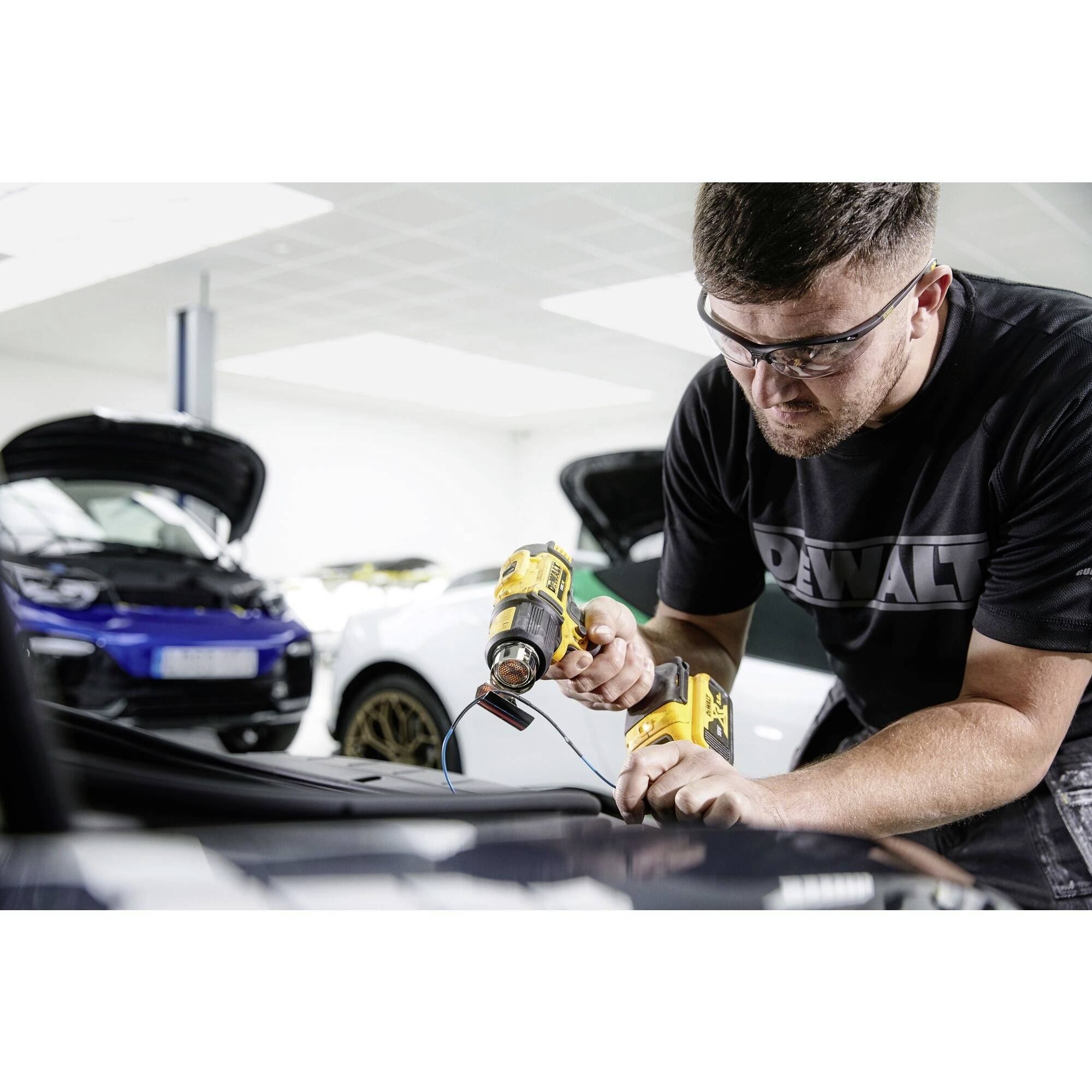 Ein Mann, der Schutzbrille trägt, benutzt ein gelbes Elektrowerkzeug zur Reparatur eines schwarzen Autos in einer Werkstatt. Im Hintergrund sind weitere Fahrzeuge sichtbar.