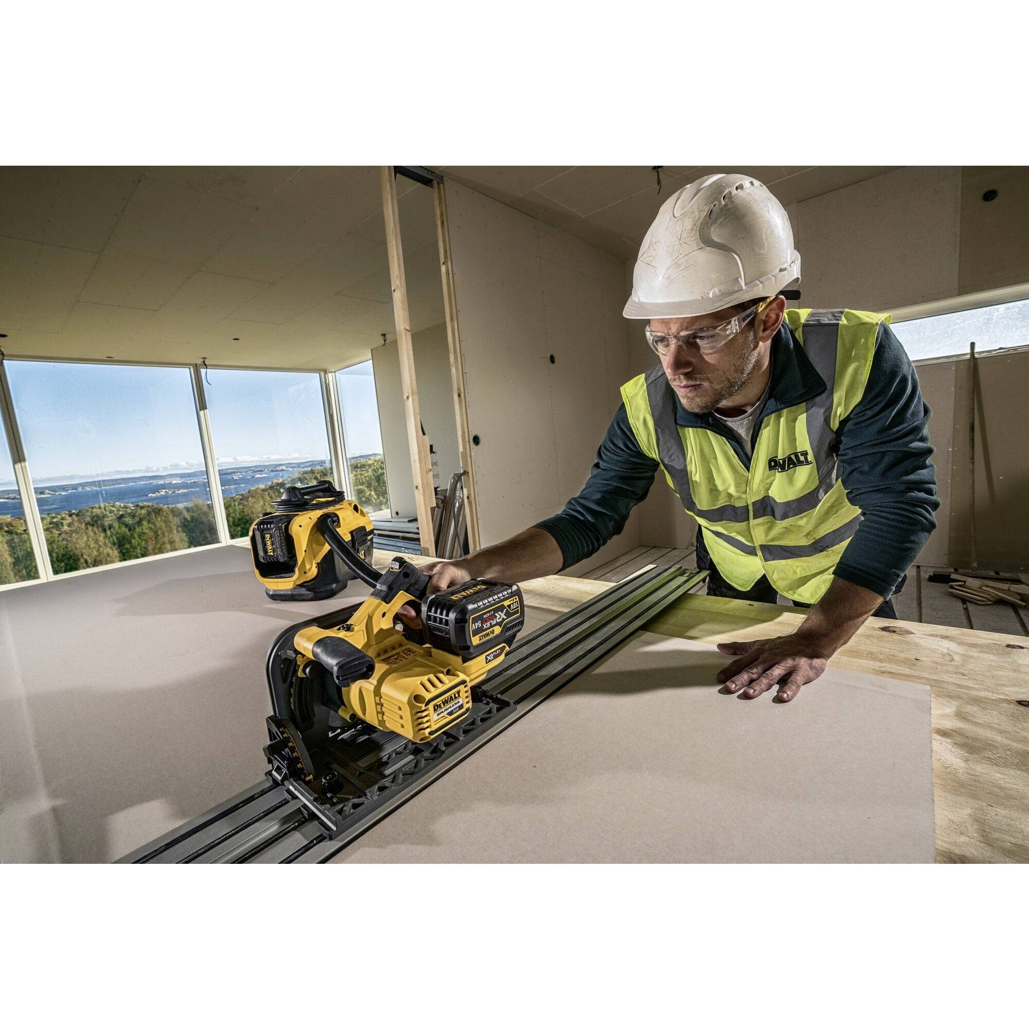 Ein Bauarbeiter mit Schutzhelm und Weste benutzt eine elektrische Säge, um Holz oder Paneele in einem Gebäude mit Panoramafenster zu schneiden.