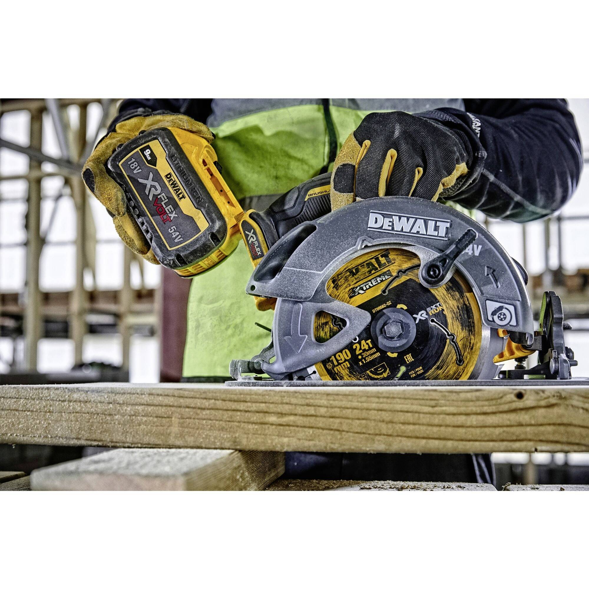 Eine Person trägt Handschuhe und nutzt eine Akku-Kreissäge der Marke DeWalt, um ein Holzbrett zu schneiden. Im Hintergrund ist ein Baugerüst sichtbar.