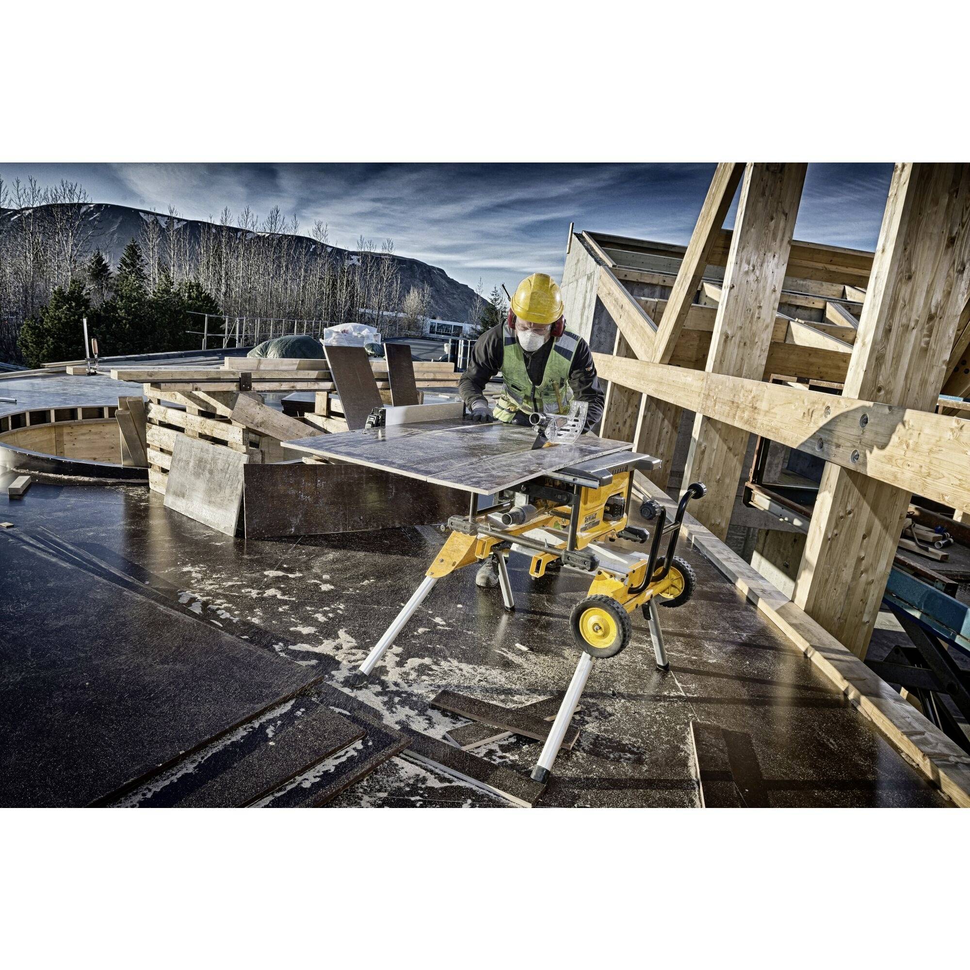 Arbeiter mit gelbem Helm und Schutzweste schneidet Holzplatte auf Baustelle. Hintergrund zeigt Berge und Baustellenstruktur.