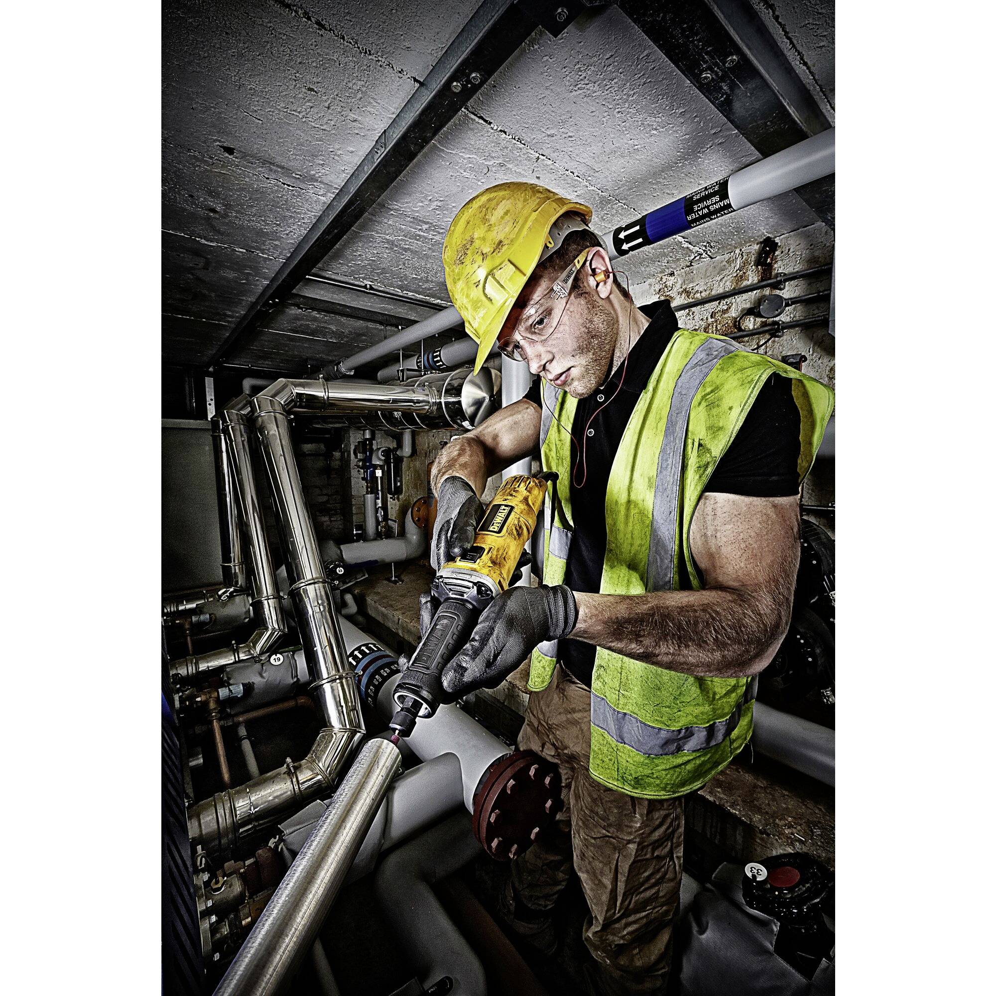 Arbeiter mit Schutzhelm und Weste repariert Rohre mit einem Werkzeug in einem industriellen Umfeld.